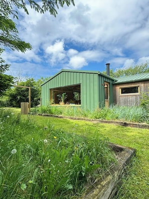 A green metal building with a large window and a chimney, surrounded by lush greenery and a clear sky.