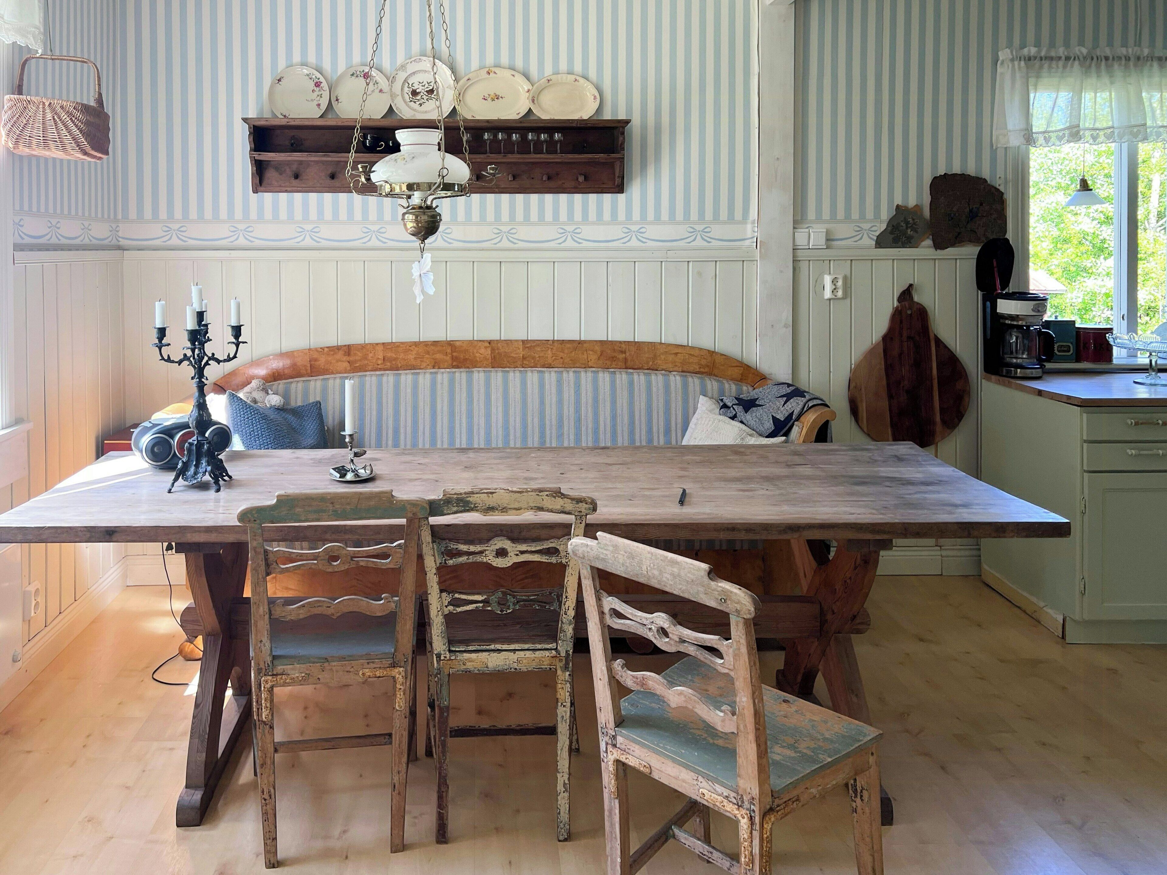 A dining area with a wooden table, two chairs, a hanging light fixture, and a wall-mounted shelf with plates.