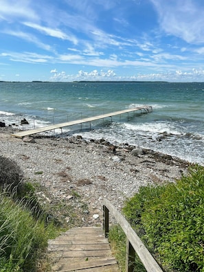 Un muelle de madera se extiende hacia una costa rocosa, con olas rompiendo sobre él.