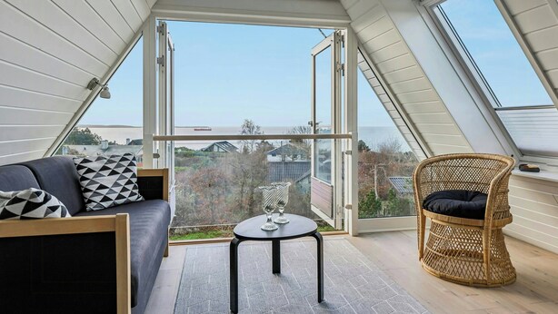 A room with a high ceiling, a sofa with cushions, a wicker chair, a small table, and large windows offering a view of the outside. - Summer house with panoramic sea views on Røsnæs (Kalundborg)