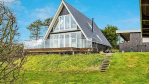A modern house with a large glass window, a wooden deck, and a grassy hill. - Summer house with panoramic sea views on Røsnæs (Kalundborg)