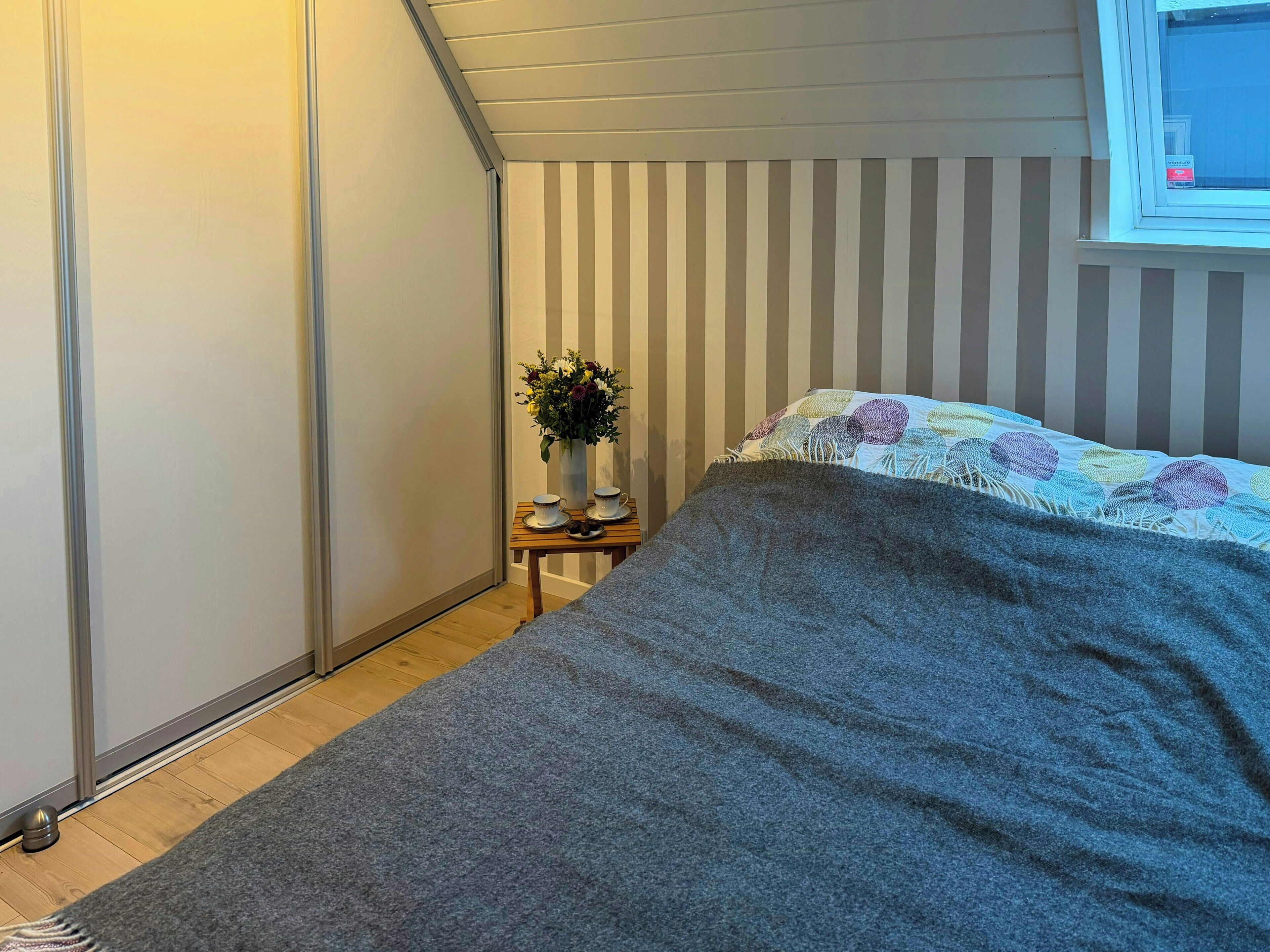 A bedroom with a bed, a small table with a vase of flowers, and a window with blinds.