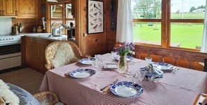 A dining area with a table set for four, wooden cabinets, a kitchen area, and a view of a grassy field through a window. - Log house near Kisserup with vintage charm (Holbæk)
