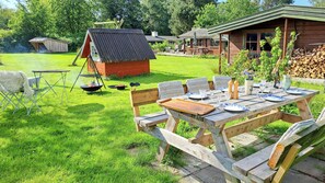 A wooden deck with a picnic table set for dining, a small barbecue grill, and a wooden shed in a grassy backyard. - Log house near Kisserup with vintage charm (Holbæk)