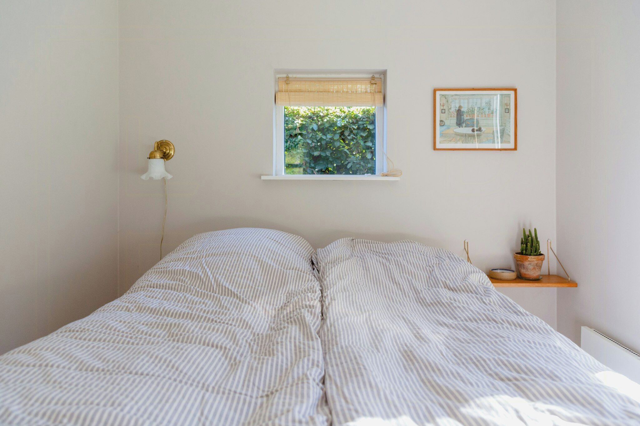 Une chambre à coucher avec un lit, une fenêtre donnant sur un paysage verdoyant, une table de chevet avec une plante et un tableau accroché au mur.