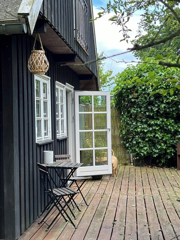 Une terrasse en bois avec une table et des chaises, une jardinière suspendue et une grande porte ouverte donnant sur un jardin.