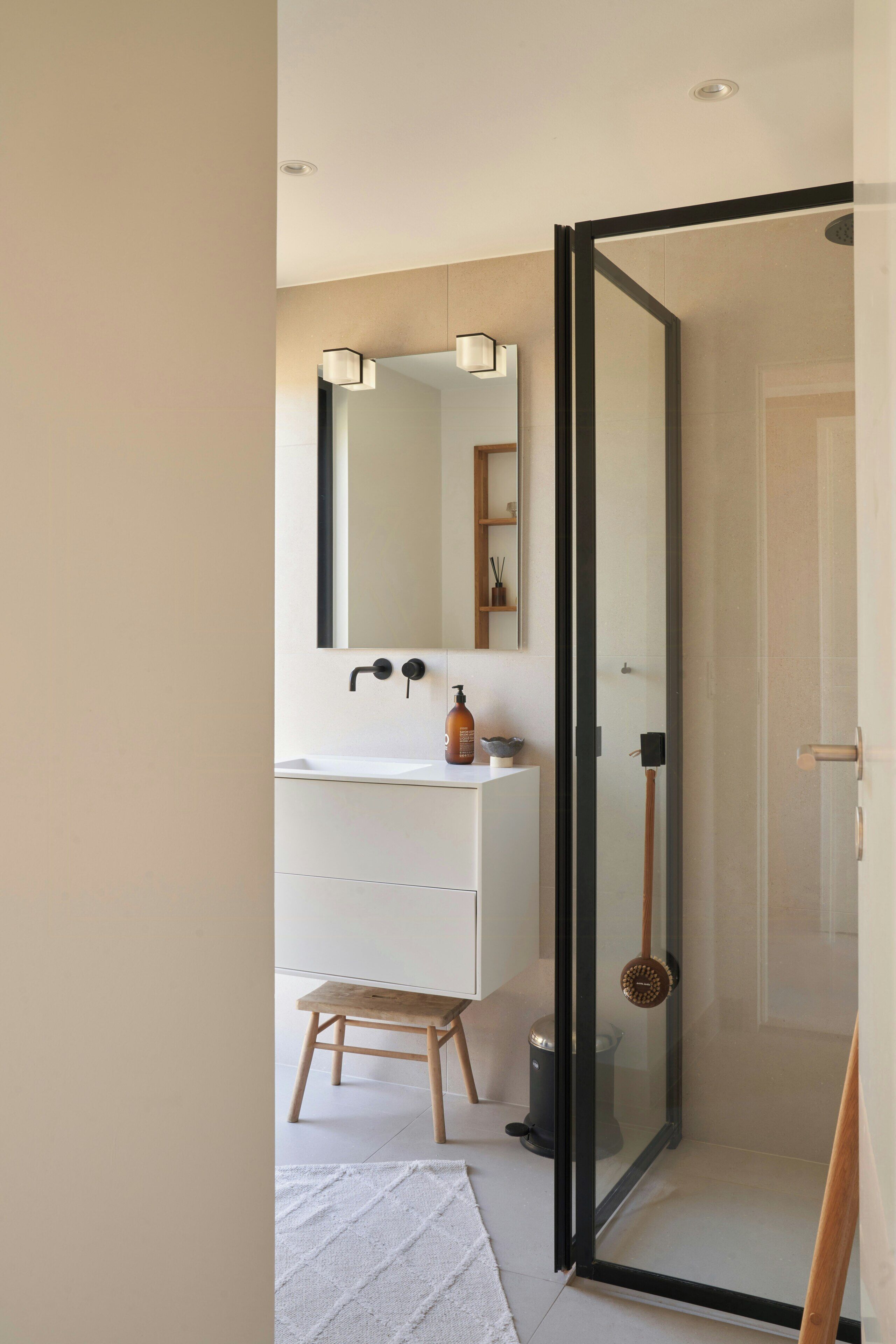 Een moderne badkamer met een douchecabine van glas, een witte wastafel en een houten krukje.
