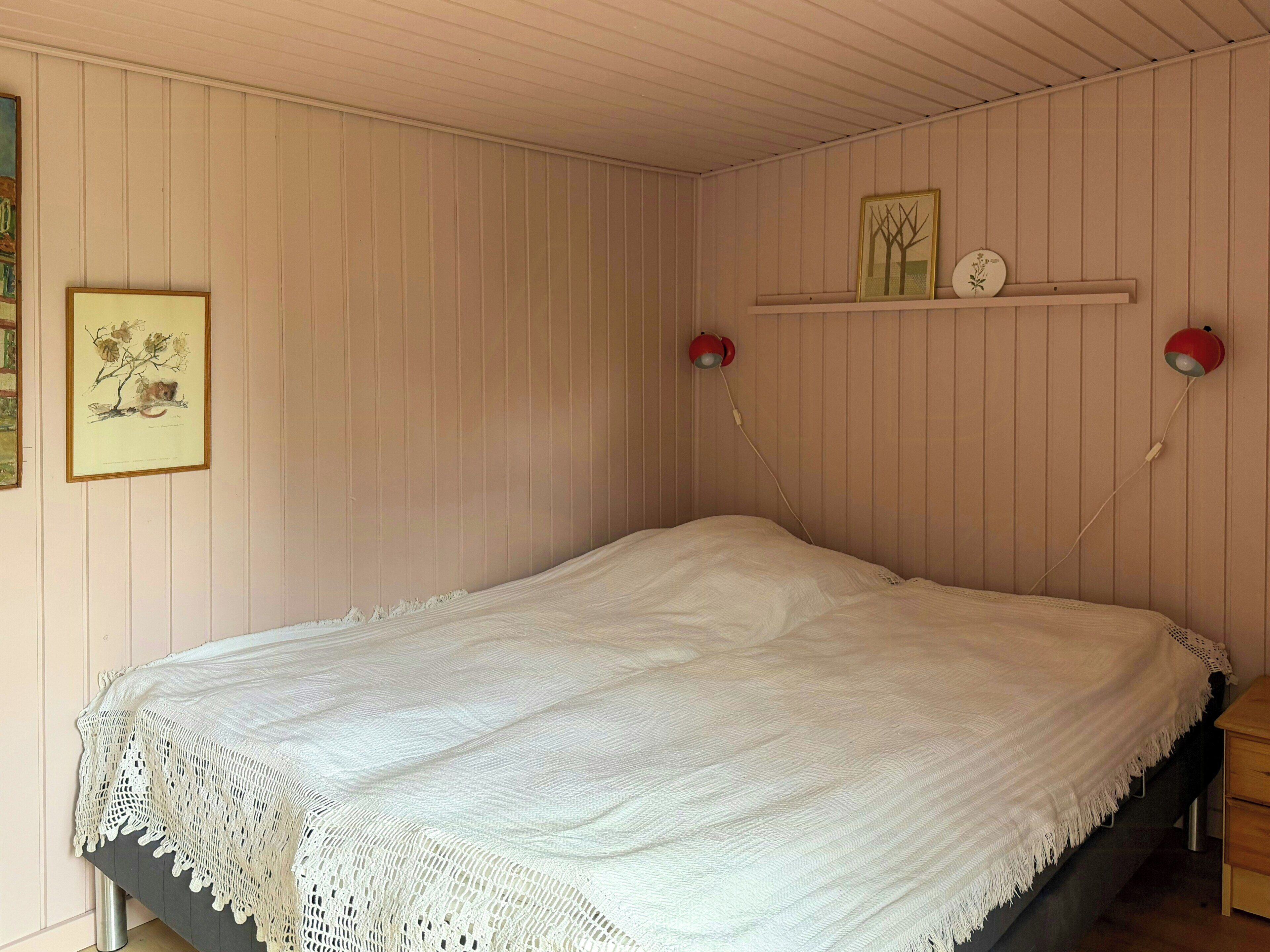 Una cama individual con una manta blanca de punto, una lámpara de pared roja y un cuadro en la pared.