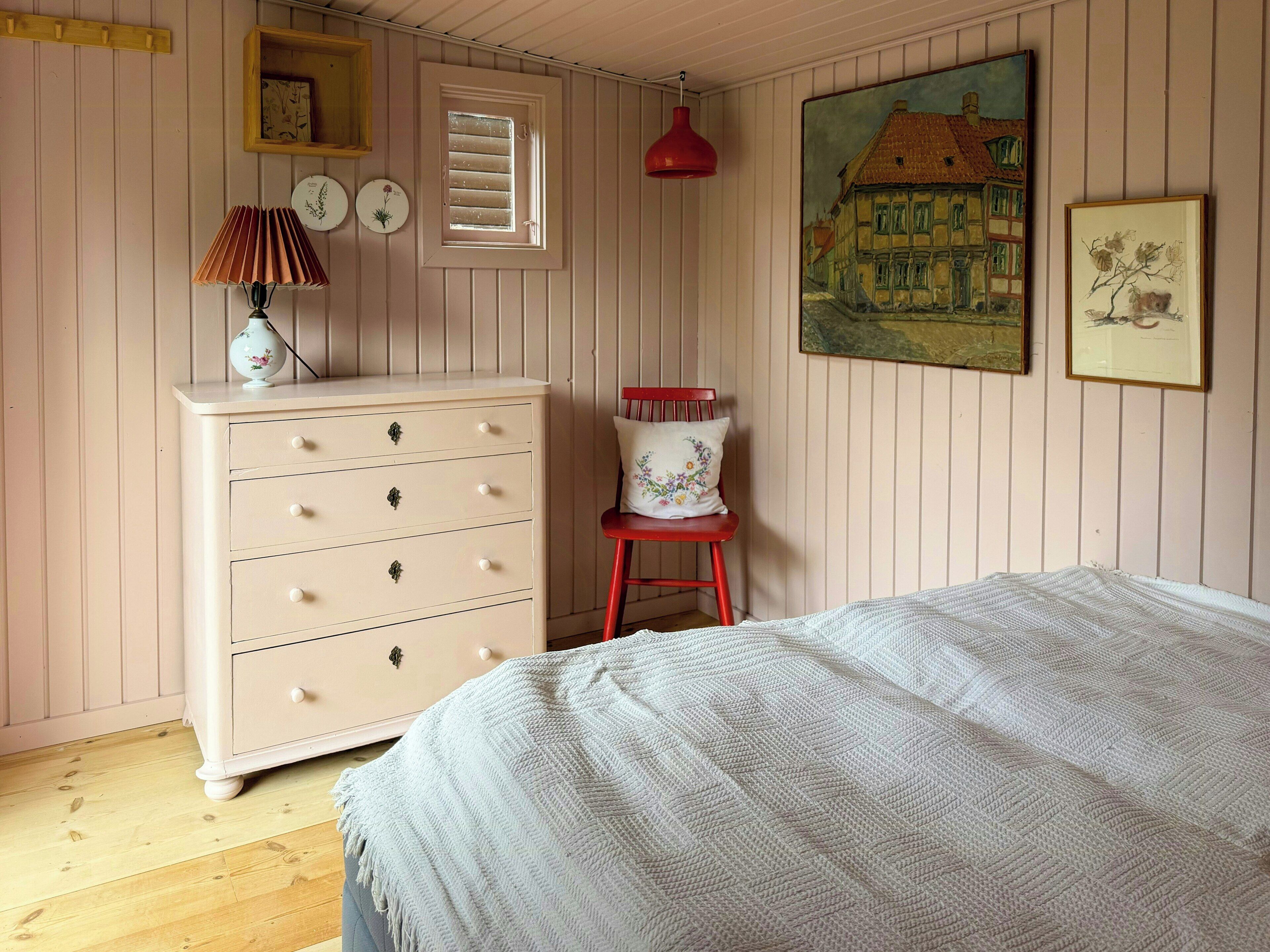 Une chambre à coucher avec un lit, une commode, une chaise, une lampe rouge et des œuvres d’art mural.