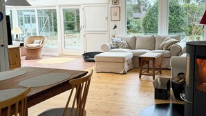 A cozy living room with a sofa, ottoman, wooden dining table, chairs, a fireplace, and large windows.