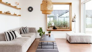 A modern living room with a sofa, a coffee table, a hanging rattan light fixture, a window with a view of greenery, and a small side table. - Newly built summer house with sea view (Haderslev)