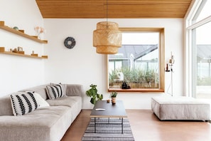 A modern living room with a sofa, a coffee table, a hanging rattan light fixture, a window with a view of greenery, and a small side table. - Newly built summer house with sea view (Haderslev)