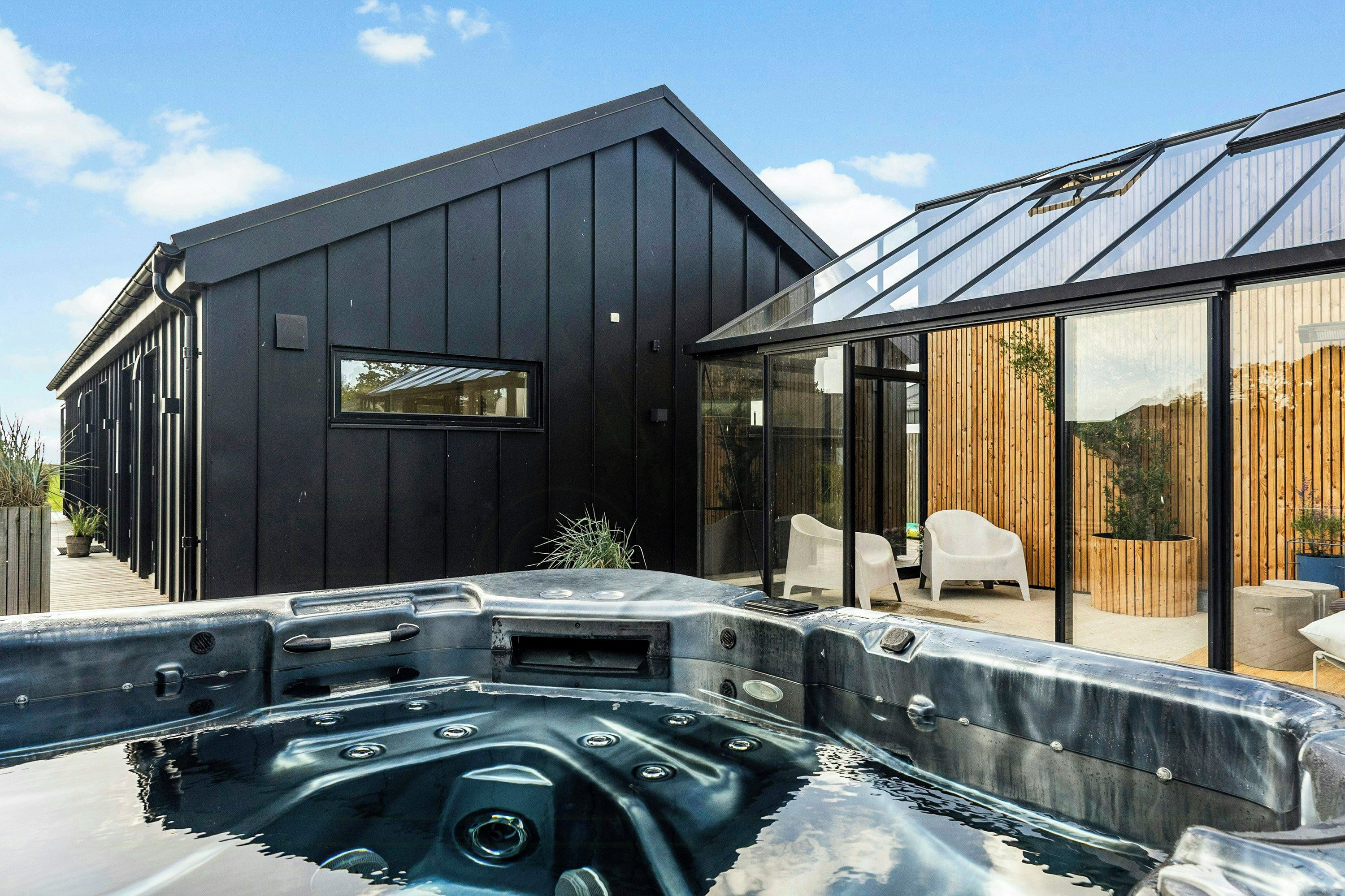A modern house with a hot tub in the foreground, a wooden deck, and a glass-enclosed patio area.