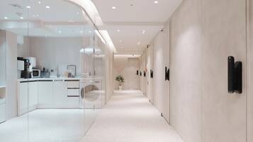 A modern hotel corridor with a curved glass wall on one side, a kitchenette, and a long hallway with doors.