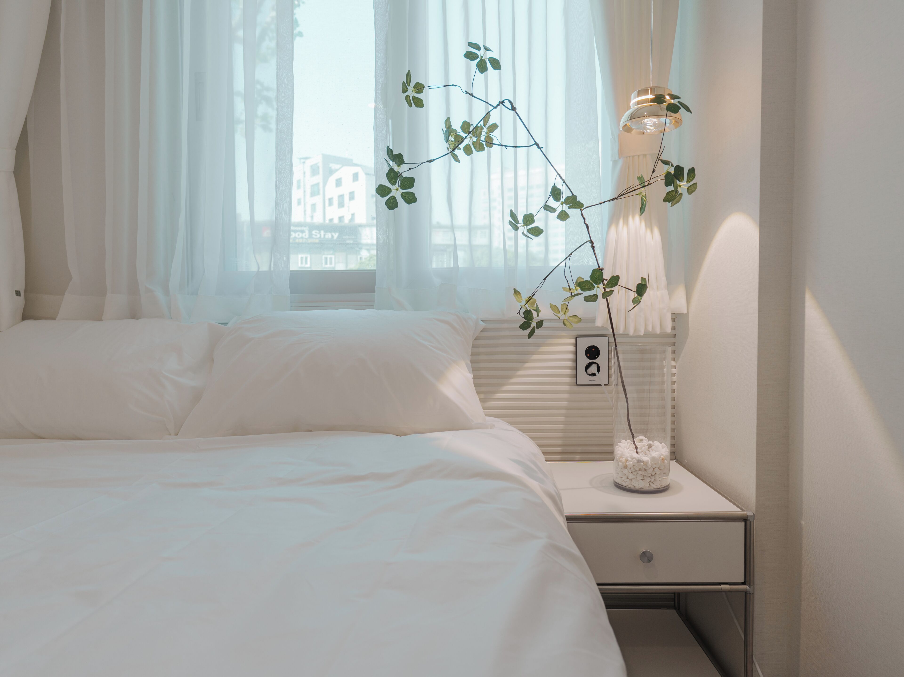 Een slaapkamer met een bed, een nachtkastje met een lamp en een raam met dunne gordijnen.