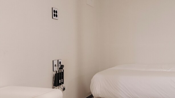 Habitación de hotel minimalista con una cama, una pequeña mesa blanca, un calentador de pared y un enchufe.