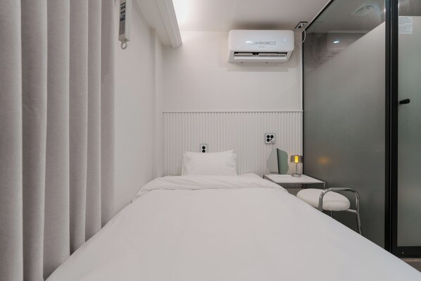 A hotel room with a bed, a bedside table with a lamp, an air conditioning unit, and a glass door.