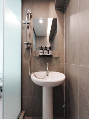 Un baño con un lavamanos pedestal blanco, un cabezal de ducha manual, un espejo sobre el lavamanos y una repisa con artículos de tocador.