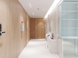 Un pasillo con puertas de madera, un dispositivo blanco fijado a la pared, un armario blanco con espejo y una mampara de ducha de vidrio.