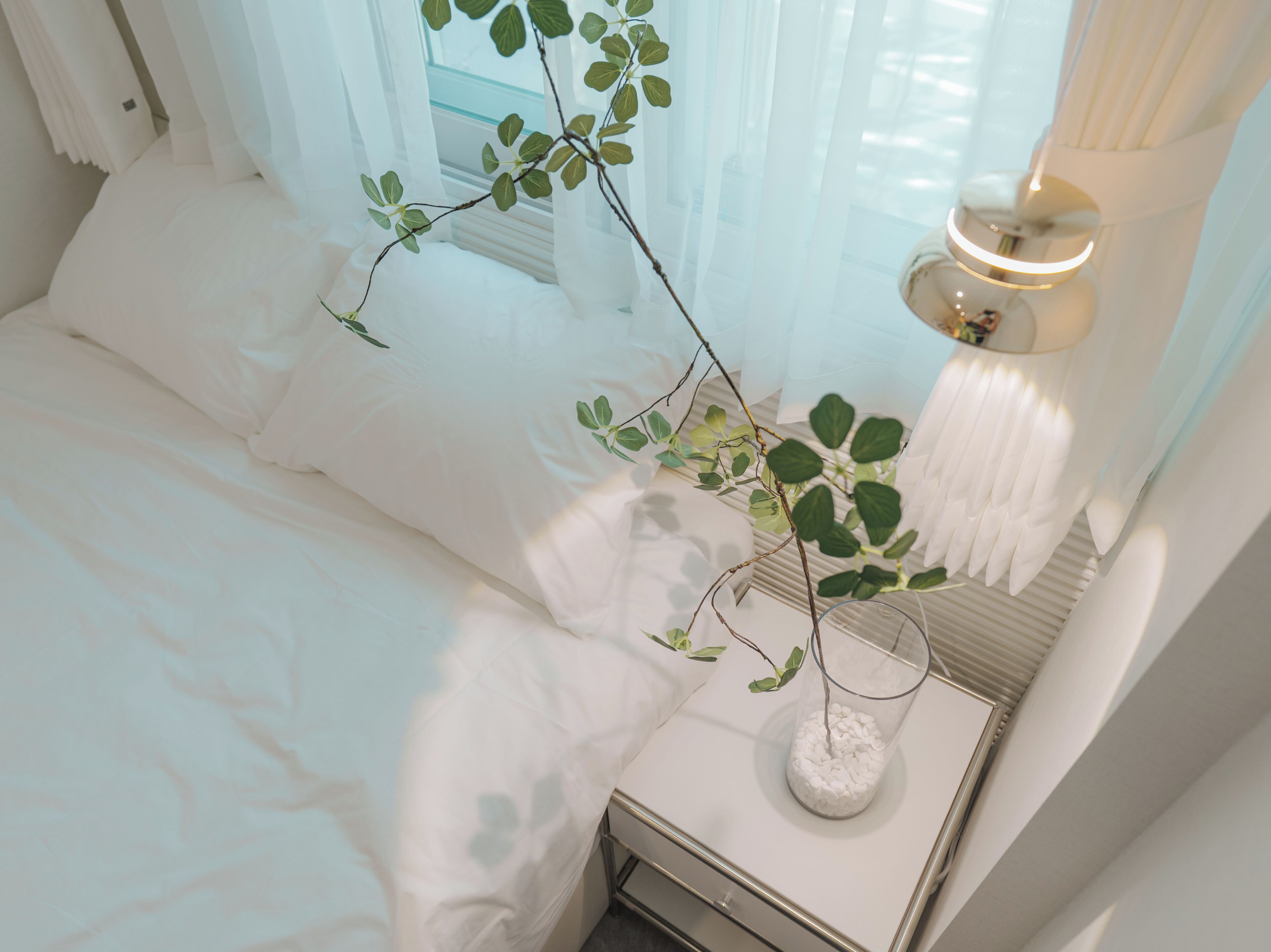 A neatly made bed with white linens, a bedside table with a glass vase containing a plant, and a lamp on the nightstand.