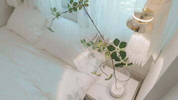 A neatly made bed with white linens, a bedside table with a glass vase containing a plant, and a lamp on the nightstand.
