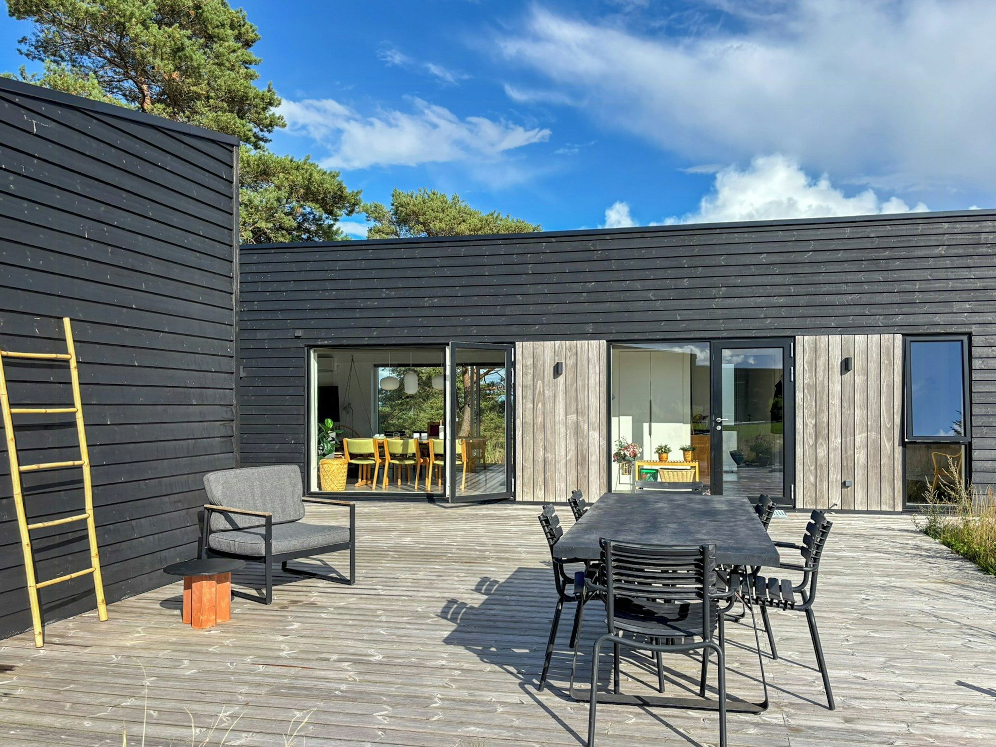 Een modern terras buiten met een houten terras, een eettafel met stoelen en een gele ladder tegen een donker houten muur.