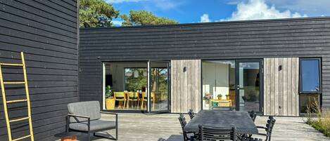 Een modern terras buiten met een houten terras, een eettafel met stoelen en een gele ladder tegen een donker houten muur.