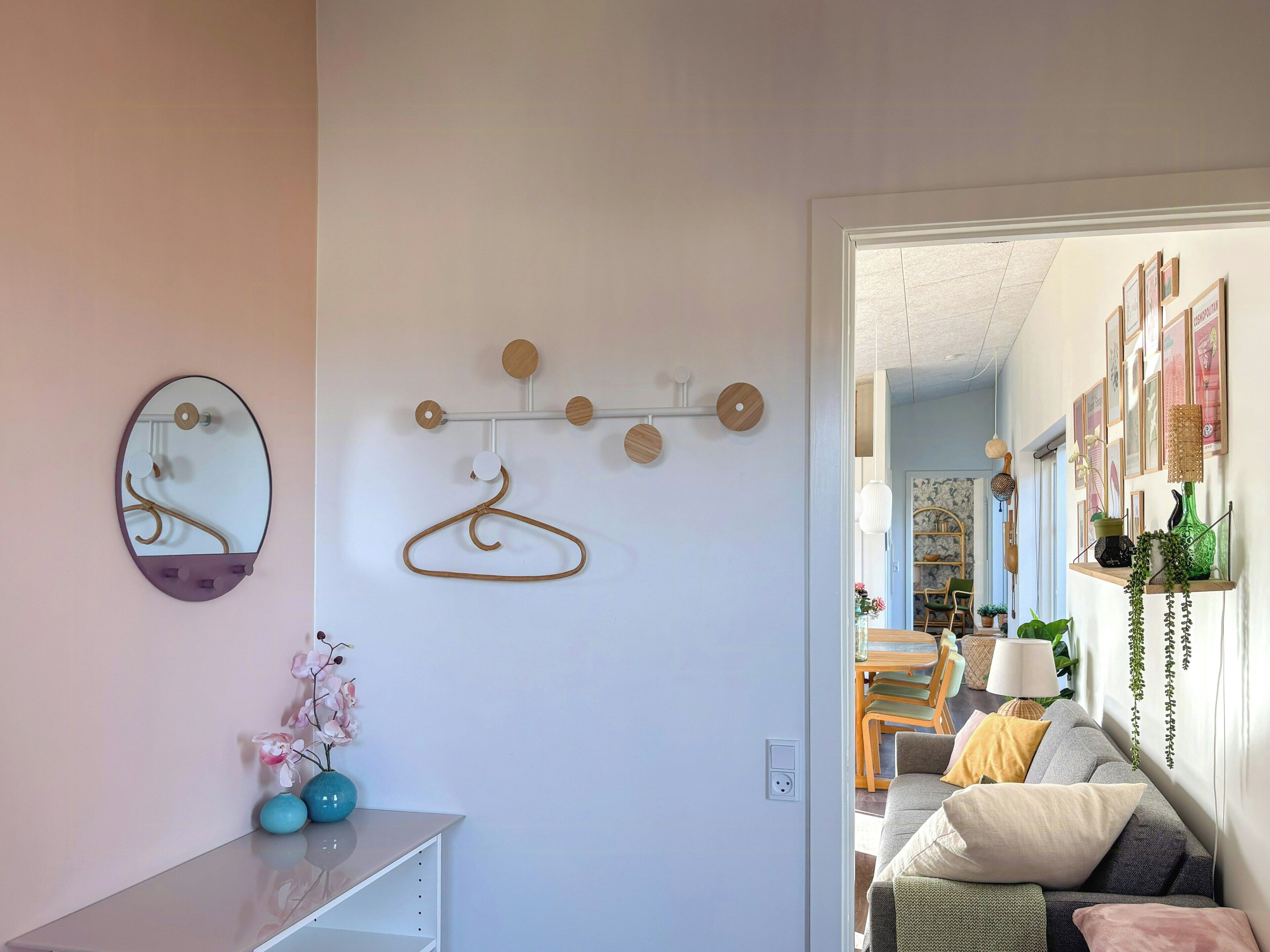 Ein Zimmer mit Spiegel, einem Wandhaken, einer Vase mit Blumen und einer Tür zu einem weiteren Raum mit Sofa und Pflanzenregal.