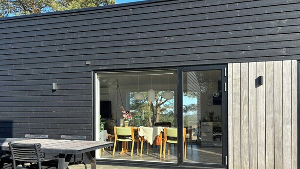 Ein modernes Gebäude mit Holzterrasse, Essbereich mit Tisch und Stühlen und einer großen Glasschiebetür zu einem Innenraum.