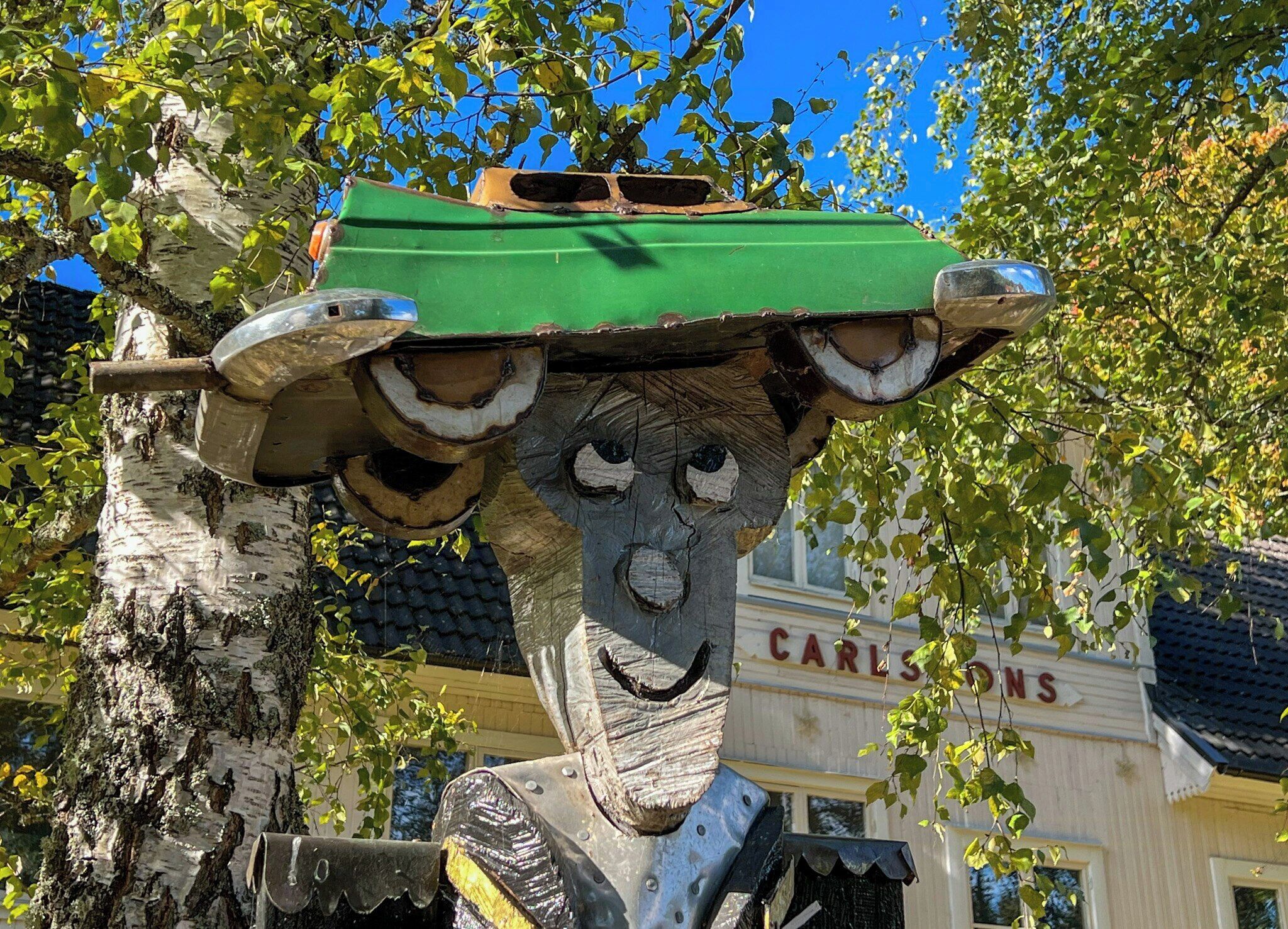 Een sculptuur met een groene hoed en gezicht, voor een gebouw met 'CARLSONS' aanduiding.