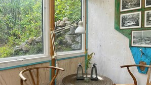 A room with a wooden chair, a small round table, a window with a view of greenery, and a wall with framed pictures. - Artist studio by Helge River in Skåne (Broby)