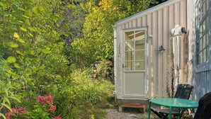 A small, single-story structure with a door and windows, surrounded by dense greenery and a gravel path. - Artist studio by Helge River in Skåne (Broby)