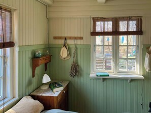Ein Zimmer mit einem Bett, einem Nachttisch mit Lampe, einem an der Wand hängenden Hut, einem Fenster mit Jalousien und einem kleinen Regal mit Büchern.