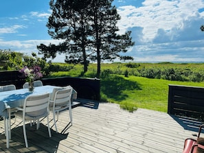 A wooden deck with outdoor furniture, a table with a vase of flowers, and a view of a grassy field and trees. - Log cabin in the front row overlooking the sea (Tranekær)
