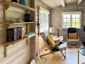A wooden cabin interior with a bookshelf, two chairs, a lamp, and a window with curtains. - Log cabin in the front row overlooking the sea (Tranekær)