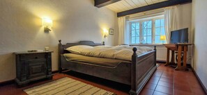 A bedroom with a bed, bedside lamps, a nightstand, a TV, and a window with curtains. - Thatched house on a Warft near Schlüttsiel (Dagebüll)