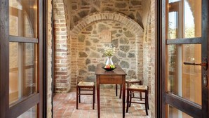 Arcada de pedra que dá acesso a uma mesa de madeira com cadeiras, um vaso com flores e uma fruteira.