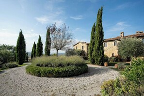 Un camino de grava que conduce a un edificio de piedra con un jardín que incluye cipreses y un seto circular.