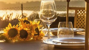Mesa posta para dois, com um copo de água, um copo de vinho e um prato de comida, com girassóis e vista para o oceano ao fundo.