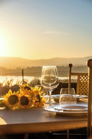 Ein Tisch für zwei Personen, gedeckt mit einem Glas Wasser, einem Weinglas und einer Speiseplatte, im Hintergrund Sonnenblumen und der Blick auf das Meer.
