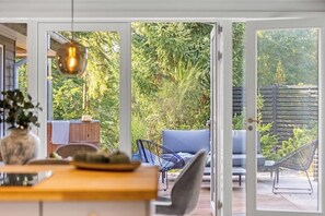 Eine geöffnete Schiebetür führt auf eine Terrasse mit einem Holztisch, Stühlen und einer hängenden Lampe.