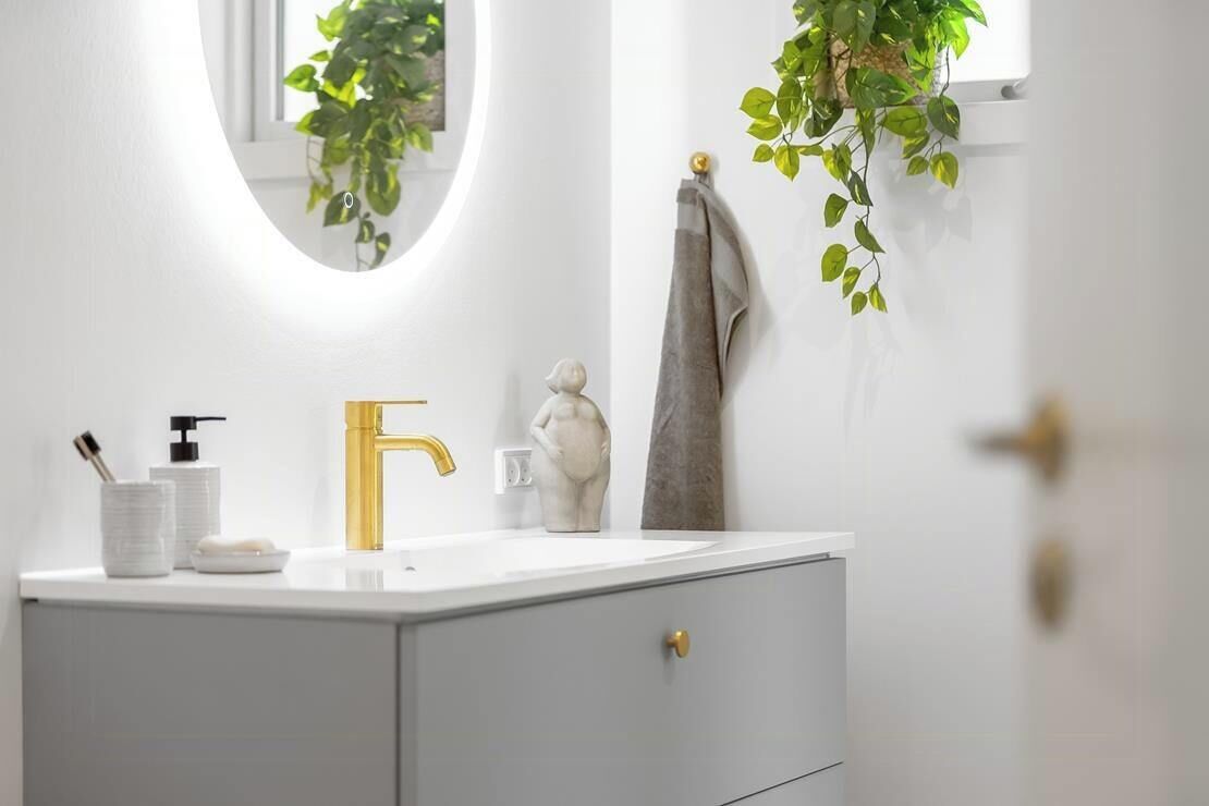 Une salle de bain avec un meuble-lavabo blanc, un robinet doré et un miroir agrémenté d’une plante.