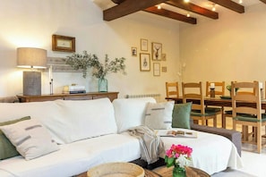 Ein gemütliches Wohnzimmer mit einer Couch, einem Esstisch und Stühlen. An der Wand hängen gerahmte Bilder, und auf dem Tisch steht eine Lampe sowie eine Vase mit Blumen.