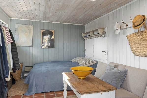Ein Zimmer mit einem Bett, einem Holztisch mit einer gelben Schüssel, einem Sofa, einem Korb und einem an der Wand befestigten Regal mit Hüten.