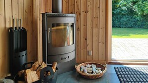A wood-burning stove, a glass table, a wicker basket with stones, and a wooden deck visible through a glass door. - Home near beach with wilderness bath (Hurup Thy)