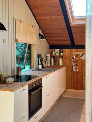A modern kitchen with a wooden ceiling, a large window, and a built-in oven. - Modern 80s holiday home with a wild garden (Fårevejle)