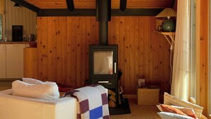 A cozy cabin interior with a wooden ceiling, a central wood-burning stove, a bed with a blanket, and a wicker basket. - Modern 80s holiday home with a wild garden (Fårevejle)