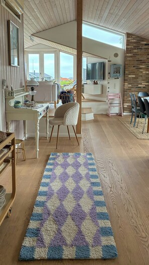 Uma sala de estar acolhedora com piso de madeira, um tapete estampado, uma mesa branca com abajur, uma cadeira e uma televisão.