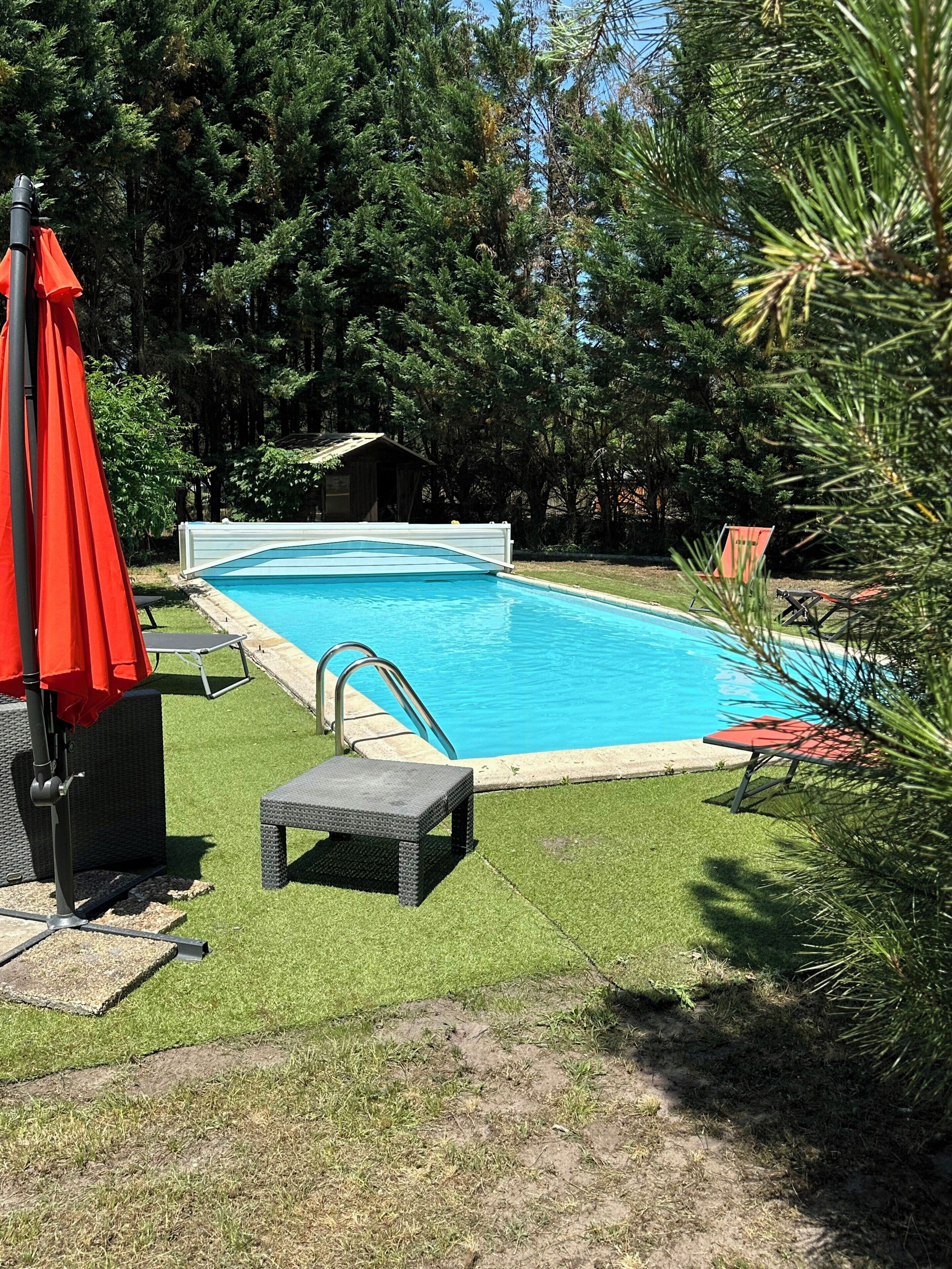 Una piscina rectangular con una escalera metálica, rodeada de césped artificial, una pequeña terraza y sombrillas rojas.