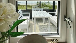 Una ventana con vistas a una terraza, una silla blanca, una mesa blanca y un jarrón con flores blancas.